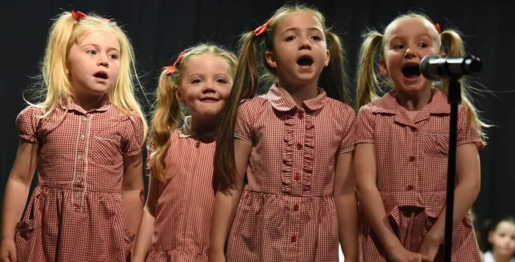 Four young children dressed in school uniforms, singing in front of a microphone